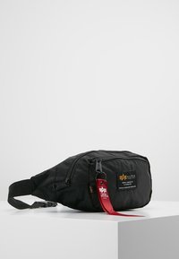 Svart midjeväska med två dragkedjefack, justerbar rem och röd tygtagg märkt "Alpha Industries USA."