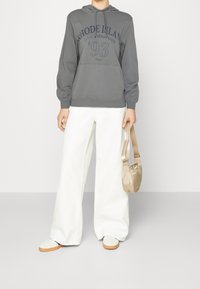 Osoba ubrana w szary hoodie z napisem "Rhode Island 93", białe szerokie spodnie, białe sneakersy i trzymająca małą beżową torebkę.