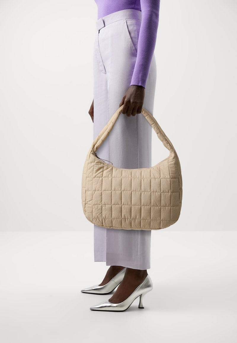 Personne tenant un sac à main matelassé beige, portant un haut à manches longues lavande, un pantalon violet clair et des chaussures à talons hauts argentées sur fond blanc.