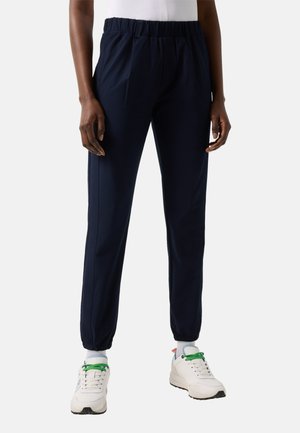 Personne portant un pantalon de jogging bleu marine à taille élastique et des baskets blanches avec des lacets verts, debout devant un fond uni.