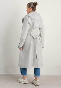 Lichtgrijze trenchcoat met een capuchon, ceintuur in de taille en lange mouwen. Heeft een split aan de achterkant en is gecombineerd met opgerolde denim en sneakers.