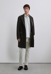 Ciemnobrązowy wełniany płaszcz z klapą z wycięciem, jednorzędowy fason, boczne kieszenie, nałożony na koszulę w kratę i jasne spodnie. Czarne buty.