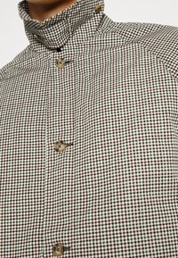 Chaqueta de botonadura delantera hecha de tela texturizada con un pequeño patrón de cuadros en blanco y negro y botones marrones. Presenta un diseño de cuello alto.