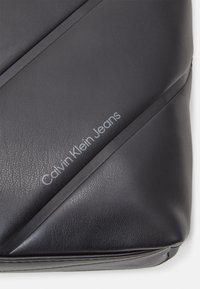Black faux leather pouch with diagonal stitch design. Features embossed "Calvin Klein Jeans" logo. Smooth texture and rectangular shape.