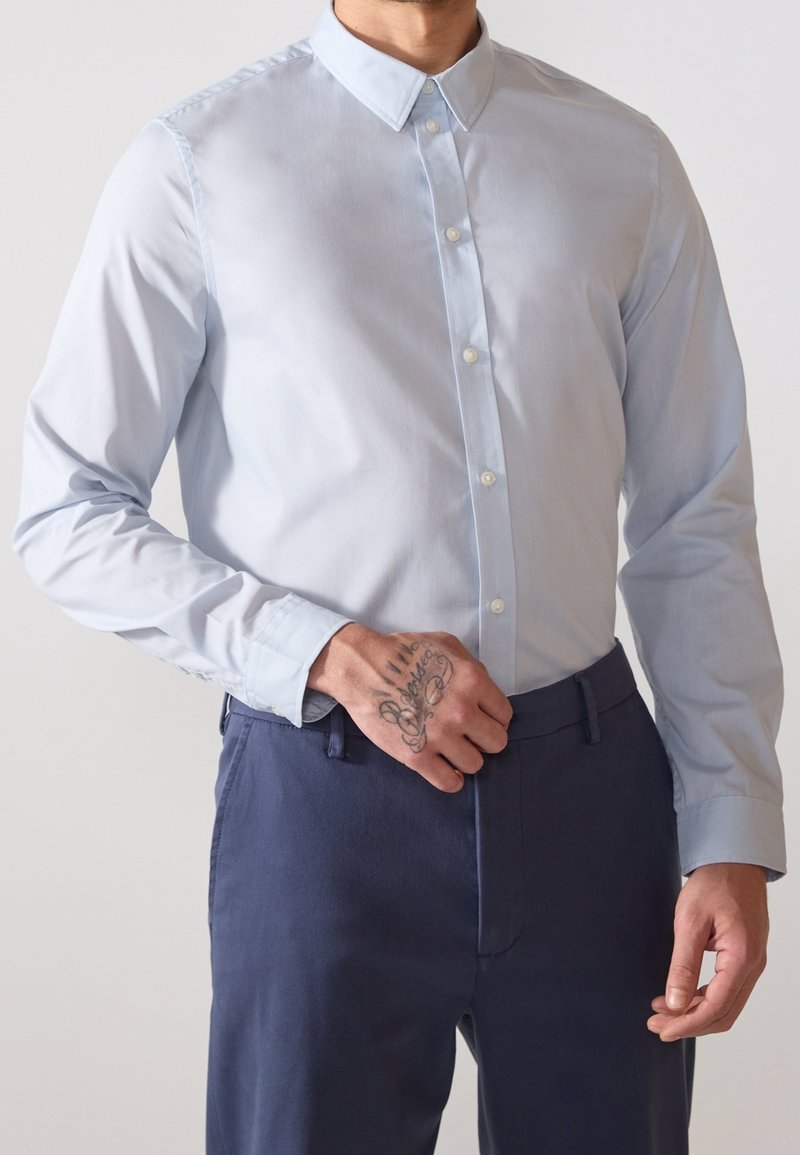 Homme portant une chemise bleu clair boutonnée et un pantalon bleu foncé, main reposant sur le pantalon montrant un tatouage sur les doigts.