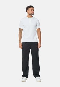 Chemise henley blanche à manches courtes avec une patte de boutonnage, associée à un pantalon noir à coupe détendue et des baskets blanches. Modèle debout, droit.