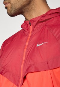 Ελαφρύ μπουφάν σε κόκκινο και πορτοκαλί με κουκούλα και πλήρες φερμουάρ, με ασημένιο λογότυπο Nike και υφή ύφασματος για αναπνευσιμότητα.
