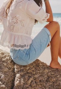 Blusa blanca bordada con mangas cortas y volantes, combinada con shorts de mezclilla azul claro. Modelo sentada sobre una superficie rocosa junto a la playa.