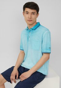 Jasnoniebieska koszulka polo z miękkiej tkaniny, z kieszenią na piersi, trzema guzikami oraz kontrastującymi granatowymi akcentami przy kołnierzyku i rękawach.