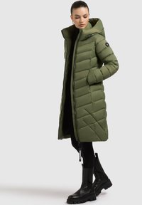 Lang olivenfarvet puffercoat med diagonal quiltning, høj krave og lynlås. Stylet med sorte leggings og kraftige sorte støvler.