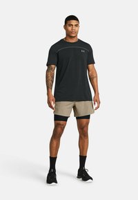 Camiseta atlética negra de manga corta, pantalones cortos beige y zapatillas negras. Presenta un tatuaje en la pierna, calcetines negros y un acento de logotipo colorido.