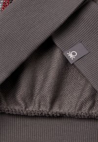 Tissu gris avec une texture côtelée et une doublure lisse. Inclus une petite étiquette en tissu avec un logo. L'ourlet présente un détail de couture froncé.