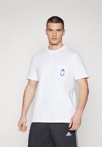 Hvit bomullt-skjorte med korte ermer, rund hals og en lomme med et blått adidas-logo. Brukt sammen med mørke shorts.