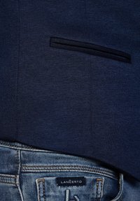 Granatowa marynarka o teksturowanym wykończeniu, z pojedynczą kieszenią na piersi, w połączeniu z wyblakłymi niebieskimi dżinsami, z widoczną metką marki.