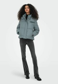 Chaqueta acolchada de color azul claro con un cuello alto, cierre delantero con cremallera y detalles de botones a presión, combinada con jeans oscuros acampanados y botas negras hasta el tobillo.