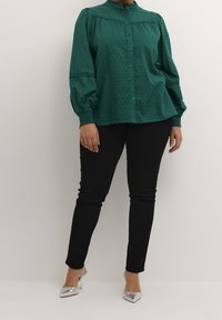Groene blouse met knopen, voorzien van gestructureerde verticale lijnen en een hoge kraag, gecombineerd met zwarte skinny jeans en zilveren puntige hakken.