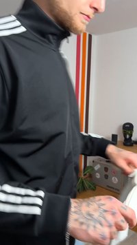 Μαύρο μπουφάν με φερμουάρ και άσπρες ρίγες, τατουάζ χέρι κρατάει ένα λευκό αντικείμενο. Το φόντο περιλαμβάνει πολύχρωμες ρίγες και διάφορα αντικείμενα πάνω σε ένα γραφείο.