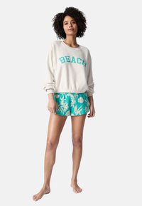 Suéter de crema claro con la palabra "BEACH" en texto turquesa, combinado con pantalones cortos florales turquesa. Textura suave, corte relajado y pantalones cortos de longitud a mitad del muslo.