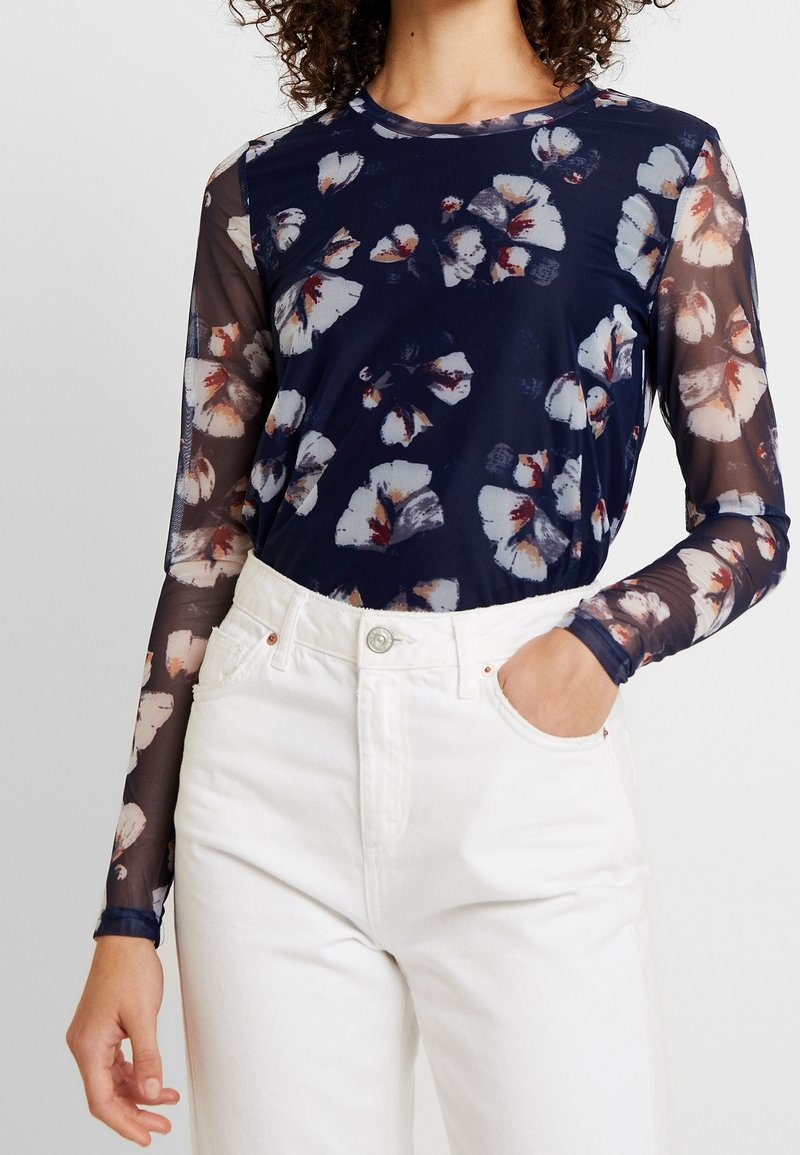 Marinblommig långärmad topp i genomskinligt material, prydd med vita och bourgogne blommor, kombinerad med högmidjade vita jeans.