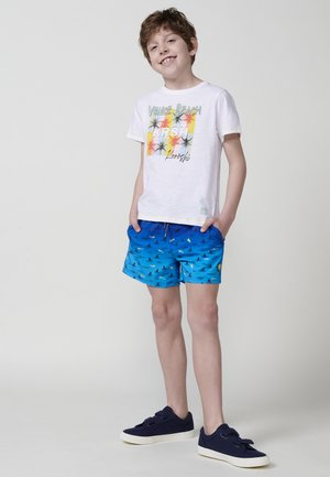Weißes T-Shirt mit buntem Palmbaum-Design, kombiniert mit blauen Badehosen mit Vogelmuster. Getragen mit navyblauen Sneakern.