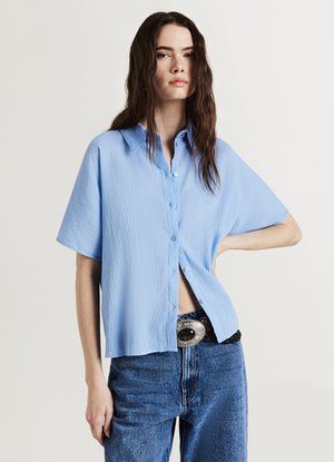 Camicia azzurro chiaro a maniche corte con bottoni, finitura testurizzata e taglio dritto, indossata con denim a vita alta e una cintura nera decorativa.