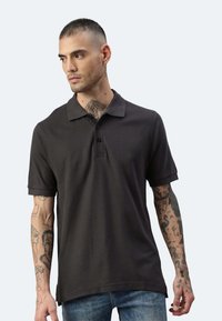 Schwarzes Polohemd mitKragen, kurzen Ärmeln und einer drei-knopfigen Leiste, getragen mit blauen Jeans. Die Arme zeigen sichtbare Tattoos.