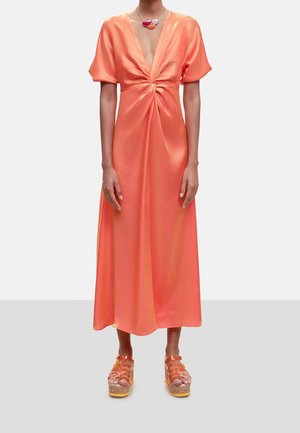 Mujer con un vestido midi de satén color coral con nudo delantero, mangas abullonadas, escote en V profundo y sandalias de plataforma naranja, de pie sobre un suelo gris.