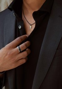 Zwarte overhemden met knopen onder een donker colbert, met een zilveren ketting met een rechthoekige hanger en een zwarte ring om de hand.