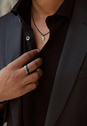Black button-up shirt under a dark suit jacket, with a silver chain necklace featuring a rectangular pendant and a black ring on the hand.