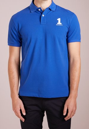 Mies, joka pukeutunut siniseen lyhythihaiseen poolopaitaan, jossa valkoinen "1 Hackett" -logo vasemmassa rinnassa, ja mustiin housuihin, seisoo yksinkertaisen taustan edessä.