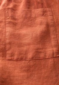 Lněná tkanina v hnědočervené barvě se šitým obdélníkovým náprsním kapsou, na které jsou viditelné stehy a textura látky.