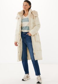 Lång krämfärgad puffjacka med fuskpälskrage, dragkedja och tryckknappar, kombinerad med blå jeans och ljusa sneakers. Mjuk, vadderad textur.