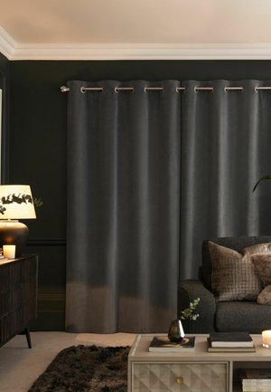 Dark grey velvet curtains with metal grommets hang in a cozy living room with a sofa, pillows, a lamp, candles, and a coffee table with books.