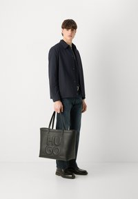Juodas odinis tote krepšys su įspaustu "HUGO" logotipu, su dviem rankenomis ir tvirtu siluetu. Nešioja stovintis modelis, apsirengęs laisvalaikio apranga.
