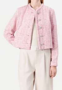 Růžový tweedový krátký kabátek s kulatým výstřihem, šesti kovovými knoflíky a strukturovanou tkaninou. Kombinovaný se světlými širokými kalhotami.