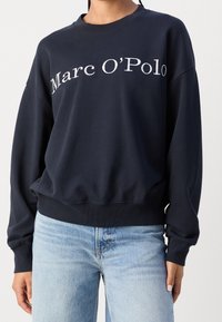 Granatowa bluza z okrągłym dekoltem, luźnym krojem i prążkowanymi mankietami. Zawiera białe logo "Marc O’Polo", wykonana z miękkiej mieszanki bawełny.