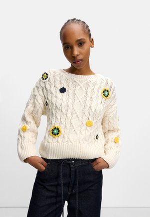 Cream cable-knit sweater with colorful floral appliqués in navy, yellow, and green. Ribbed cuffs and hem. Paired with dark denim jeans.