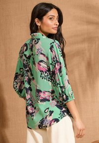 Žalia bliuska su abstrakčiomis rožinėmis ir juodomis dekoracijomis, trijų ketvirčių ilgio rankovėmis ir raukšlėtu apykakle. Lengvas audinys, laisvas kirpimas.