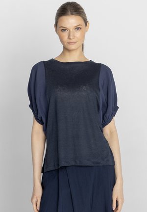 Kobieta o brązowych włosach związanych w niski kucyk, ubrana w luźny granatowy top z bufkami na rękawach i pasującą spódnicę na jednolitym tle.