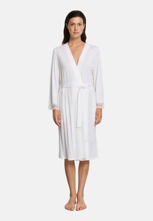 Kvinde stående barfodet iført en knælængde hvid kjortel med ærmer kantet med blonder og et bundet bælte om livet mod en ensfarvet baggrund.