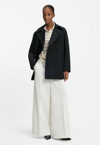 Czarny dwurzędowy płaszcz z szerokimi klapami, noszony na beżowo-kremowej pasiastym polo, w połączeniu z szerokimi białymi spodniami i czarnymi spiczastymi butami.