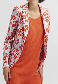 Blazer floral bleu avec des fleurs rouges et orange, associé à une robe orange texturée. Le blazer présente une coupe ajustée et un revers.