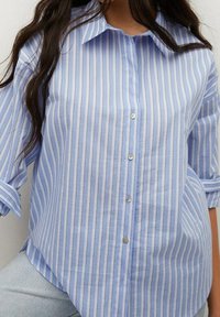 Blue and white striped button-up shirt with a pointed collar, feature loose sleeves, and a longer back hemline. Material appears lightweight.