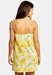 Floral jurk in het geel met een losse pasvorm, gebroken witte accenten, verstelbare strikjes op de schouders en een gegolfde zoom. Lichtgewicht stof.