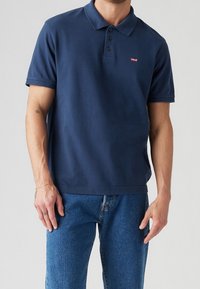 Camisa polo azul marino de algodón con una botonera de tres botones y un pequeño logo rojo de Levi's en el pecho, que complementa unos vaqueros azules informales.