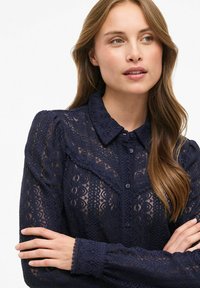 Blonde med blonde i marineblå med krave, knaplukning og strukturerede blomster kvinder. Den har pufærmer og delikate bølgede manchetter.