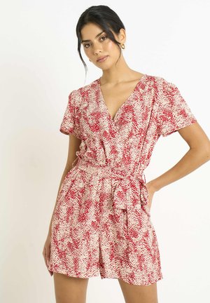 Woman wearing a red and white patterned short-sleeve romper with a V-neck and a tied waist, posing against a plain light background.