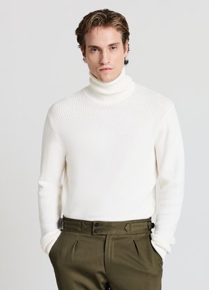 Uomo con pelle chiara e capelli ricci che indossa un maglione a collo alto bianco con texture e pantaloni verde oliva a vita alta, mani in tasca.