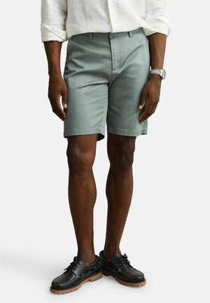 Mann iført lysegrønne shorts, hvit skjorte med lange ermer og oppbrettede mansjetter, svarte skinnsko og sølvfarget armbåndsur.