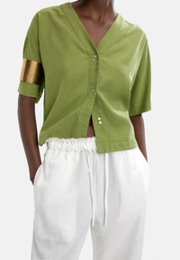 Zielona bawełniana koszula z krótkim rękawem i dekoltem w kształcie litery V, z guzikami z przodu, bocznymi rozcięciami i złotym akcentem na jednym rękawie. Połączona z białymi spodniami z elastycznym pasem.
